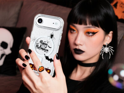 A woman with Halloween makeup holding a "Happy Halloween" themed phone case, spider earrings, and a pumpkin ring